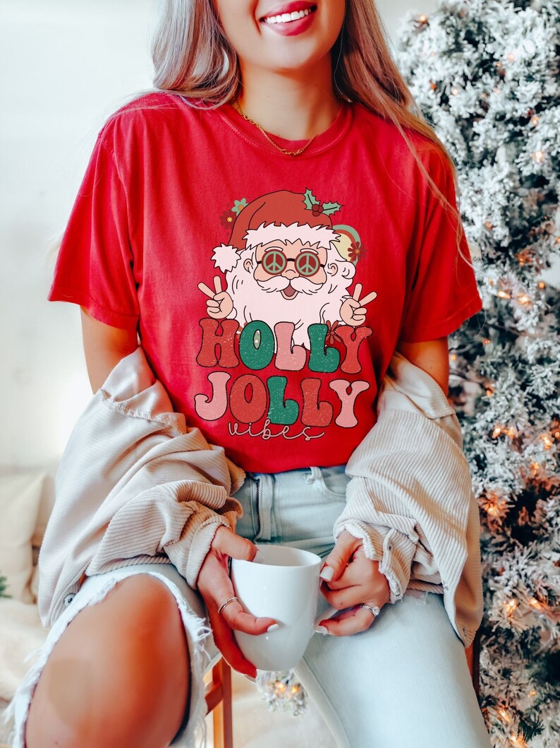 May include: Red t-shirt with a retro-style illustration of Santa Claus wearing sunglasses and peace signs. The text "HOLLY JOLLY vibes" is printed in colorful letters.