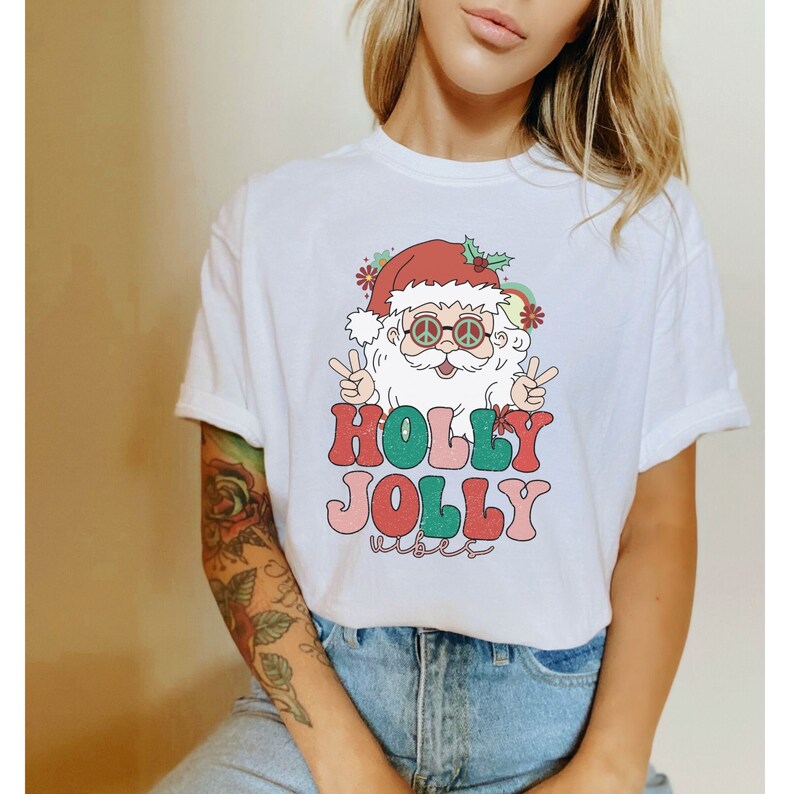 May include: White t-shirt with a retro-style illustration of Santa Claus wearing peace sign sunglasses and the text "HOLLY JOLLY vibes".