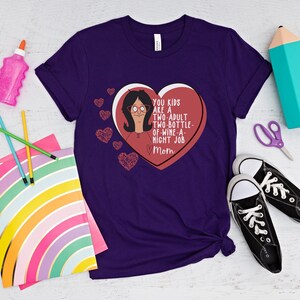 K&ouml;nnte beinhalten: Ein lila T-Shirt mit einem herzf&ouml;rmigen Aufdruck, der eine Cartoon-Frau und den Text "YOU KIDS ARE A TWO-ADULT TWO-BOTTLE-OF-WINE-A-NIGHT JOB Mom" zeigt. Das Shirt liegt neben Schulmaterial und schwarzen Turnschuhen.