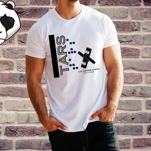 Op de afbeelding: Wit T-shirt met een zwart-wit grafisch ontwerp met de tekst "TARS" en een gestileerd robotontwerp. De tekst "U.S. MARINE CORPS Tactical Robot" is ook op het shirt te vinden.