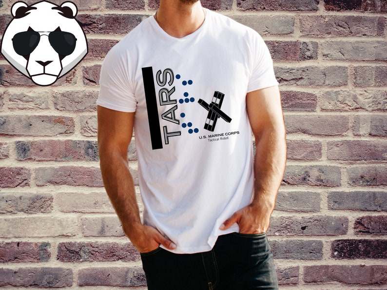Op de afbeelding: Wit T-shirt met een zwart-wit grafisch ontwerp met de tekst "TARS" en een gestileerd robotontwerp. De tekst "U.S. MARINE CORPS Tactical Robot" is ook op het shirt te vinden.