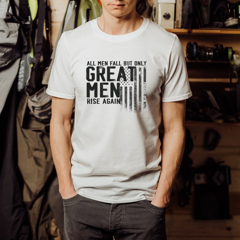 Puede incluir: Camiseta blanca con un estampado gr&aacute;fico en negro y gris de una bandera estadounidense y el texto "All men fall but only great men rise again".