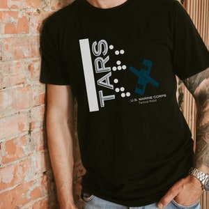 Op de afbeelding: Een zwart T-shirt met een wit grafisch ontwerp met de tekst "TARS" met een gestileerd robotontwerp en de tekst "U.S. MARINE CORPS Tactical Robot".