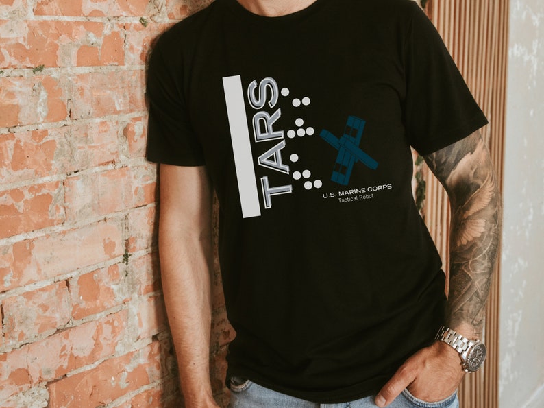 Op de afbeelding: Een zwart T-shirt met een wit grafisch ontwerp met de tekst "TARS" met een gestileerd robotontwerp en de tekst "U.S. MARINE CORPS Tactical Robot".