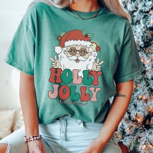 May include: A green t-shirt with a retro-style illustration of Santa Claus wearing peace sign sunglasses and the text "Holly Jolly Vibes".