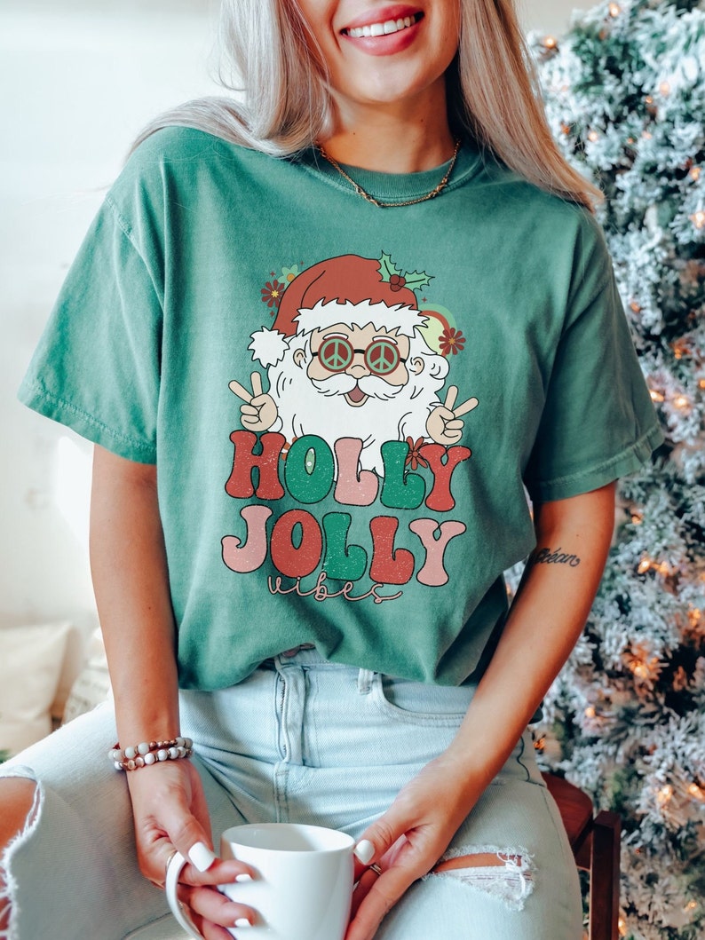 May include: A green t-shirt with a retro-style illustration of Santa Claus wearing peace sign sunglasses and the text "Holly Jolly Vibes".