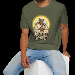 May include: Olive green t-shirt featuring a graphic of St. Brother Teresa with a golden halo. The image shows her with hands clasped in prayer, and the text "ST. BROTHER TERESA" is below the image. The person is wearing blue jeans.