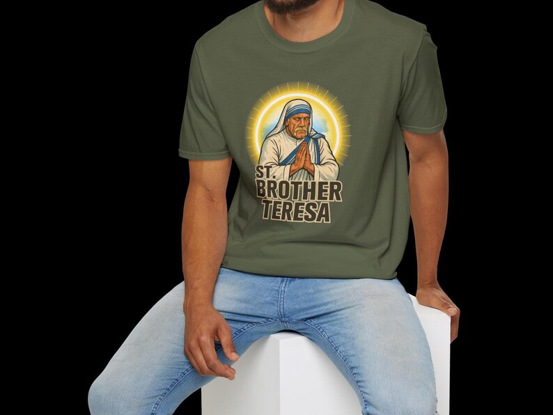 May include: Olive green t-shirt featuring a graphic of St. Brother Teresa with a golden halo. The image shows her with hands clasped in prayer, and the text "ST. BROTHER TERESA" is below the image. The person is wearing blue jeans.