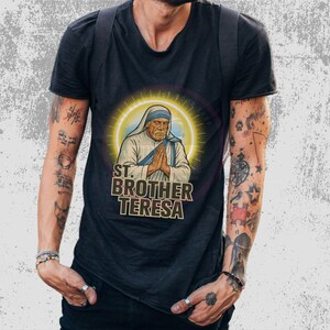 May include: Black t-shirt featuring a graphic of Mother Teresa with the text "ST. BROTHER TERESA" in a stylized font. The image of Mother Teresa is surrounded by a golden halo. The shirt is worn by a person with tattoos.