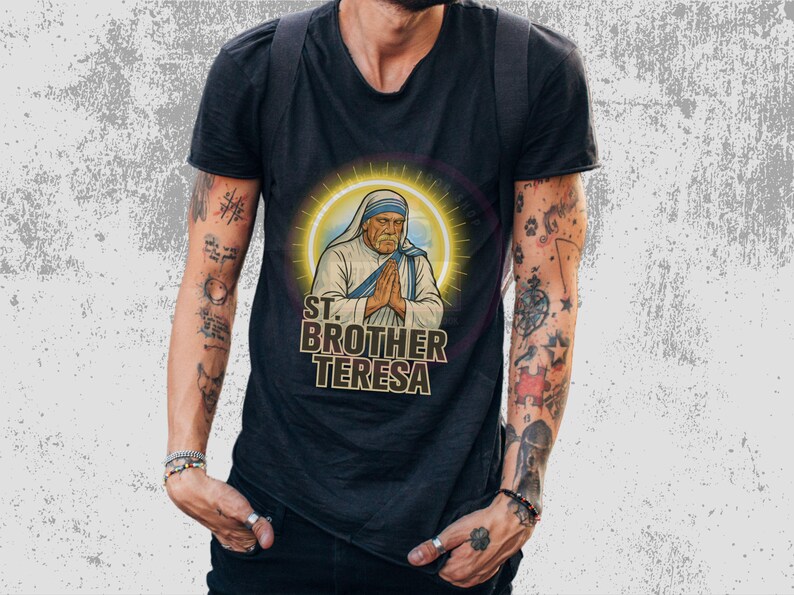 May include: Black t-shirt featuring a graphic of Mother Teresa with the text "ST. BROTHER TERESA" in a stylized font. The image of Mother Teresa is surrounded by a golden halo. The shirt is worn by a person with tattoos.