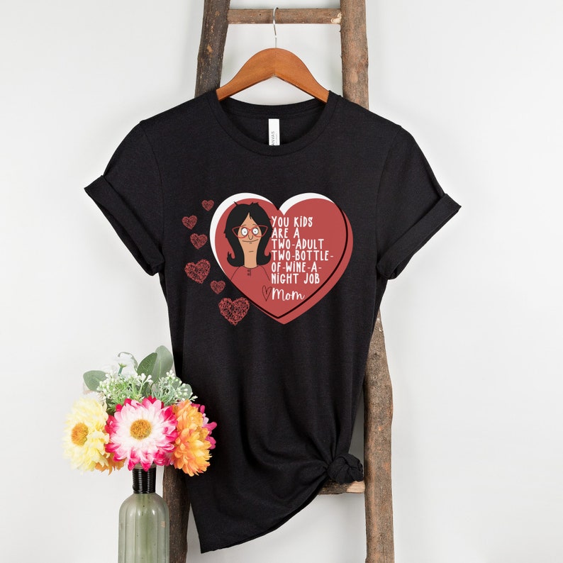 K&ouml;nnte beinhalten: Schwarzes T-Shirt mit einem herzf&ouml;rmigen Design. Das Design enth&auml;lt eine Cartoon-Figur und den Text: "YOU KIDS ARE A TWO-ADULT TWO-BOTTLE-OF-WINE-A-NIGHT JOB Mom". Mehrere kleine Herzen sind ebenfalls enthalten.
