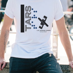 Op de afbeelding: Wit T-shirt met een zwart en grijs grafisch ontwerp. De grafiek bevat de tekst "TARS" en een gestileerd robotontwerp. De tekst "U.S. MARINE CORPS Tactical Robot" is ook inbegrepen.