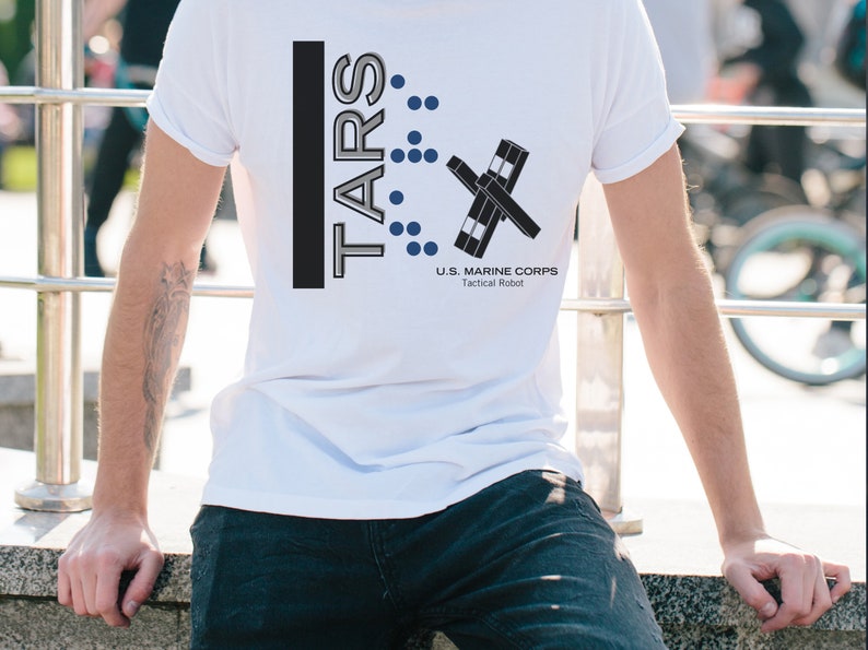 Op de afbeelding: Wit T-shirt met een zwart en grijs grafisch ontwerp. De grafiek bevat de tekst "TARS" en een gestileerd robotontwerp. De tekst "U.S. MARINE CORPS Tactical Robot" is ook inbegrepen.