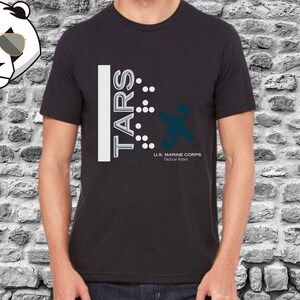 Op de afbeelding: Zwart T-shirt met een wit grafisch ontwerp met de tekst "TARS" met een reeks stippen en een gestileerd robotontwerp. De tekst onder de robot luidt "U.S. MARINE CORPS Tactical Robot".