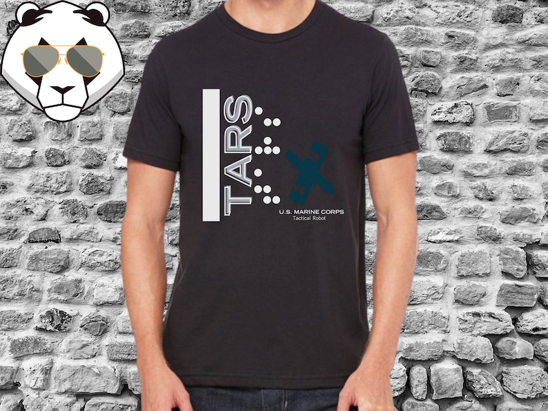 Op de afbeelding: Zwart T-shirt met een wit grafisch ontwerp met de tekst "TARS" met een reeks stippen en een gestileerd robotontwerp. De tekst onder de robot luidt "U.S. MARINE CORPS Tactical Robot".