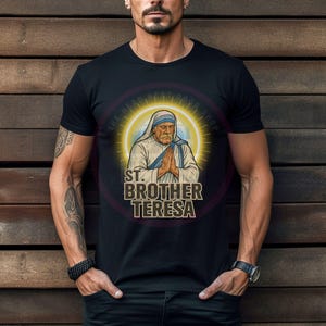 May include: Black t-shirt featuring a graphic of St. Brother Teresa with a halo and clasped hands. The text "ST. BROTHER TERESA" is displayed below the image in a stylized font. The shirt is worn by a person.