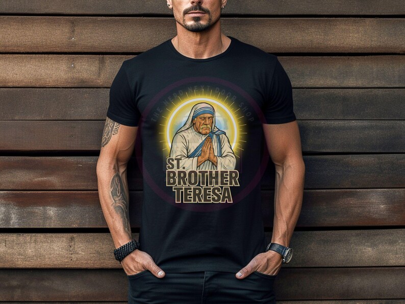 May include: Black t-shirt featuring a graphic of St. Brother Teresa with a halo and clasped hands. The text "ST. BROTHER TERESA" is displayed below the image in a stylized font. The shirt is worn by a person.