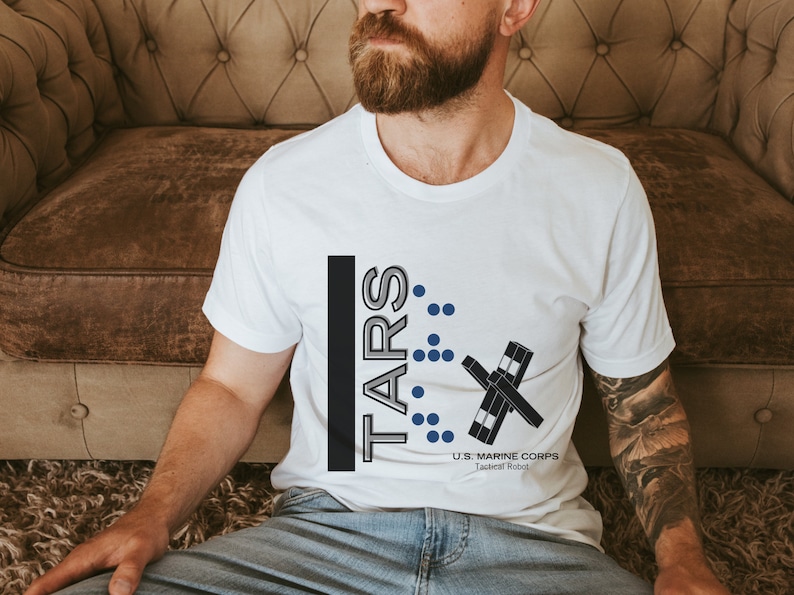 May include: A white t-shirt with a black and gray graphic design that says "TARS" with a stylized robot design and the text "U.S. MARINE CORPS Tactical Robot".