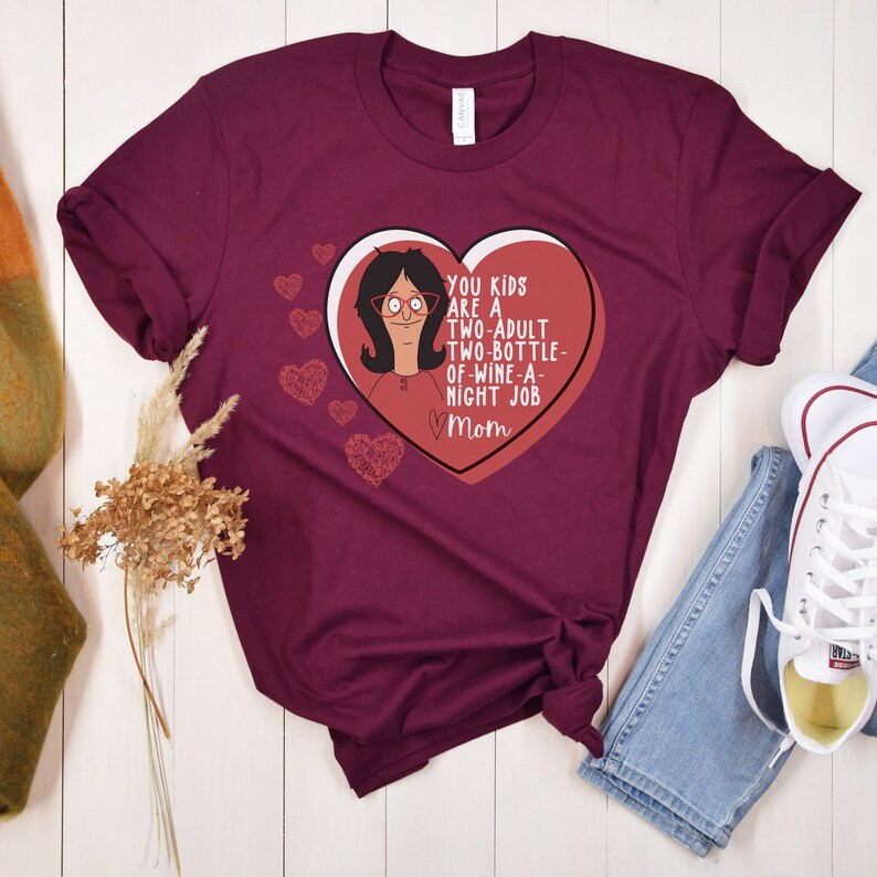 K&ouml;nnte beinhalten: Burgunderfarbenes T-Shirt mit herzf&ouml;rmiger Grafik. Die Grafik zeigt eine Zeichentrickfigur und den Text "YOU KIDS ARE A TWO-ADULT TWO-BOTTLE-OF-WINE-A-NIGHT JOB Mom". Zus&auml;tzliche Herzgrafiken sind verstreut.