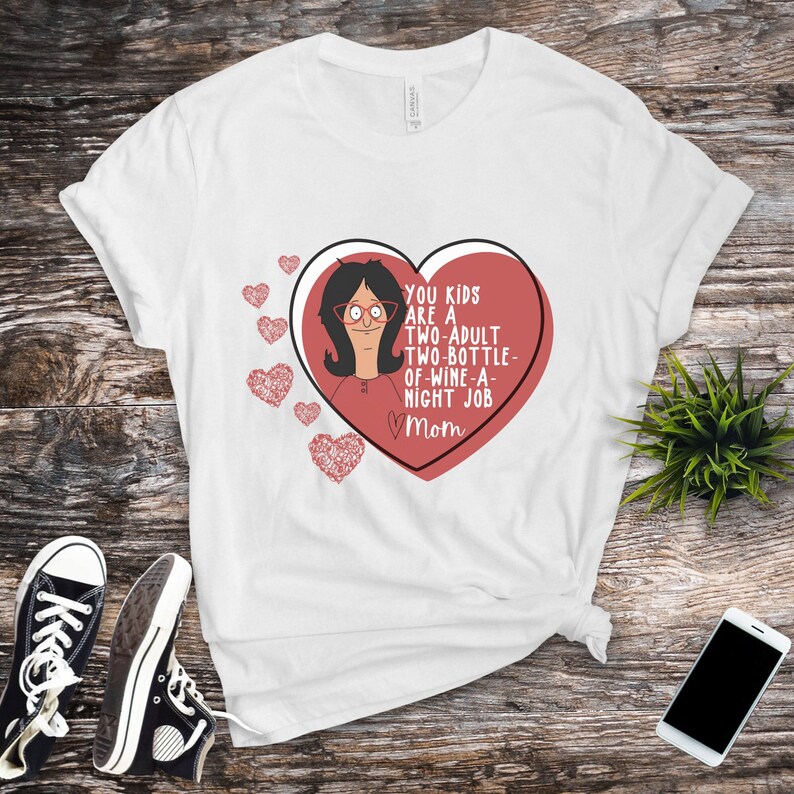 K&ouml;nnte beinhalten: Wei&szlig;es T-Shirt mit einem roten Herz-Grafik, das eine Cartoon-Frau und den Text "YOU KIDS ARE A TWO-ADULT TWO-BOTTLE-OF-WINE-A-NIGHT JOB Mom" zeigt. Das Shirt wird mit schwarzen Turnschuhen und einem Smartphone kombiniert.