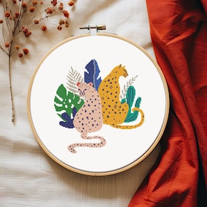 May include: Cross-stitch embroidery in a wooden hoop featuring two leopards with spots, surrounded by green, blue, and gray leaves. One leopard is pink, the other is yellow. The background is white.