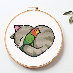 Puede incluir: Un diseño de bordado de punto de cruz que presenta un gato gris enroscado en forma de corazón, con un pájaro verde y rojo anidado en su pelaje.