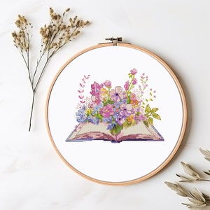 Peut inclure: Un motif de broderie au point de croix repr&eacute;sentant un livre ouvert avec un bouquet de fleurs roses, violettes et blanches. Le livre est sur un fond blanc.