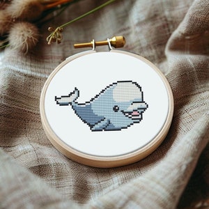 May include: A white and gray whale cross-stitch pattern in a wooden embroidery hoop. The whale is smiling and has a light blue tail.