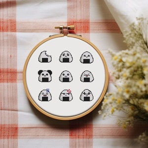May include: A white embroidery hoop with a cross-stitch design of nine different onigiri, or Japanese rice balls, with various facial expressions. The onigiri are gray with black seaweed and white details.
