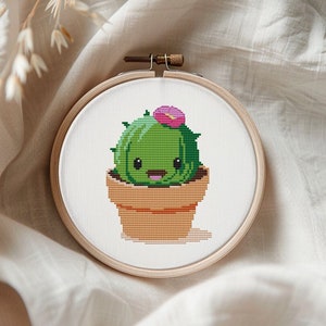Könnte beinhalten: Ein Kreuzstich-Stickrahmen mit einem niedlichen Cartoon-Kaktus in einem Topf. Der Kaktus ist grün mit einer rosa Blüte oben und einem lächelnden Gesicht.