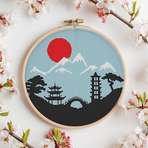 May include: A cross-stitch embroidery hoop with a design of a Japanese landscape. The design features a red sun, mountains, a pagoda, a bridge, and trees. The background is a light blue color.