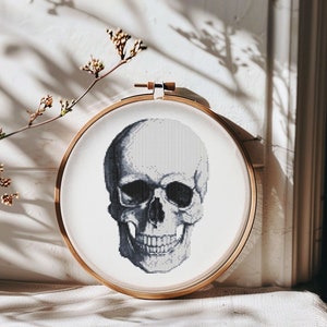 May include: A black and white cross-stitch embroidery of a human skull in a wooden embroidery hoop.