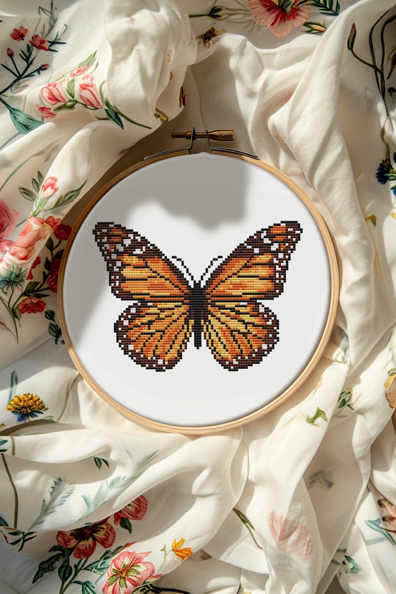May include: A close-up of a cross-stitch embroidery hoop with a monarch butterfly design. The butterfly is orange, black, and white, and is stitched on white fabric.