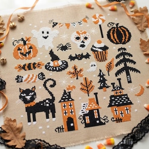 May include: A cross-stitch sampler on tan fabric featuring Halloween motifs including a black cat, jack-o'-lanterns, a ghost, a skull, a witch hat, houses, trees, a cupcake, and a lollipop, all rendered in black, orange, and white thread.