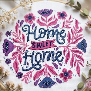 Peut inclure: Une broderie au point de croix sur tissu blanc avec le texte Home Sweet Home en bleu foncé et rose. Le motif est entouré d'une couronne florale composée de fleurs et de feuilles violettes, roses et bleues, le tout dans un cercle à broder en bois.