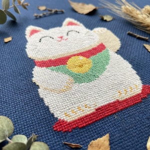 Könnte beinhalten: Eine Kreuzstichstickerei auf dunkelblauem Stoff, die eine weiße winkende Katze mit rotem Halsband, grünem Lätzchen und gelber Glocke zeigt. Die Katze hat geschlossene Augen, eine kleine rosa Nase und eine erhobene Pfote. Das Motiv ist in Weiß, Rot, Grün und Gelb gestickt.