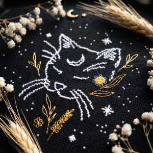 May include: Black cross-stitch design featuring a cat's face with white thread, surrounded by gold and white floral accents. The design includes a crescent moon and stars. The background is black.