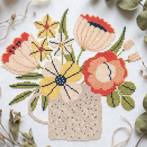 Op de afbeelding: Een kruissteek borduurpatroon op witte stof met een boeket gestileerde bloemen in tinten geel, perzik en rood, geschikt in een gespikkelde beige kan met een handvat, geaccentueerd door groene bladeren en stengels.