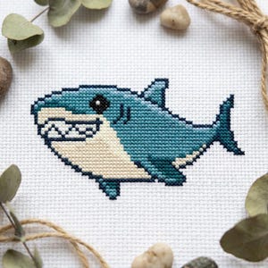 May include: A cross-stitch embroidery pattern on white fabric featuring a cartoon shark. The shark is rendered in shades of teal and blue with a cream-colored underbelly, a black eye, and a wide mouth displaying a row of white, jagged teeth.