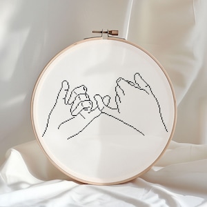 May include: A black and white line drawing of two hands intertwined, symbolizing friendship or love, on a white canvas in a wooden embroidery hoop.