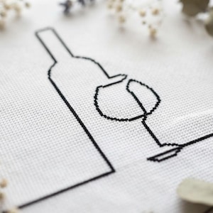May include: A white fabric piece featuring a black cross-stitch embroidery design of a wine bottle and a wine glass. The embroidery is rendered in a simple, continuous line style, creating a minimalist outline of the bottle and glass.