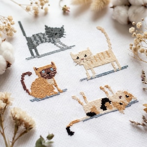 May include: A close-up of a white fabric featuring four cross-stitched cat designs. The cats are in various poses and colors, including gray, tan, and brown, with blue and black accents. The image is surrounded by dried flowers and cotton.