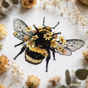May include: A cross-stitch embroidery on white fabric featuring a detailed bumblebee. The bee has a black and yellow striped body adorned with small yellow flowers. Its wings are rendered in shades of gray with intricate stitching details.
