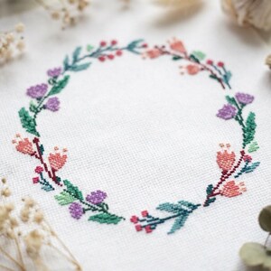 Könnte beinhalten: Ein Kreuzstich-Stickmuster auf weißem Stoff, das einen kreisförmigen Kranz aus Blumen und Blättern zeigt. Das Design besteht aus kleinen rosa, violetten und korallenfarbenen Blüten, die mit dunkelgrünem Blattwerk und Stielen in einem Ring angeordnet sind.