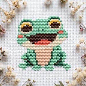 Puede incluir: Diseño de rana en punto de cruz, sonriendo, en tonos verdes, marrones y melocotón. La rana está sobre un fondo de tela blanca, rodeada de flores secas. El detallado punto de cruz crea un ambiente alegre y artesanal.
