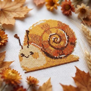 May include: A cross-stitch design of a smiling snail with a shell in shades of orange, yellow, and brown. The snail is surrounded by autumn leaves and flowers, creating a seasonal theme. The cross-stitch is on white fabric.