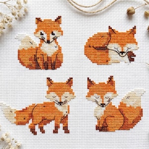 Peut inclure: Un motif de broderie au point de croix sur tissu Aida blanc représentant quatre renards orange et blancs distincts dans diverses postures, assis, endormis et debout, réalisés avec des contours noirs détaillés et des fils colorés.