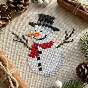 May include: A cross-stitch embroidery on beige fabric featuring a snowman with a black top hat, an orange carrot nose, a red scarf, and black twig-like arms. The snowman has three black buttons down its centre and a curved black mouth.