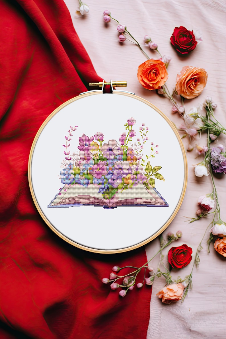 Peut inclure: Un cercle de broderie blanc avec un motif de point de croix d'un livre ouvert avec un bouquet de fleurs violettes, roses et jaunes. Le cercle est sur un fond de tissu rose et blanc avec des fleurs roses et rouges.