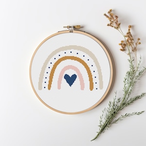 Può includere: Un cerchio da ricamo a punto croce con un design a arcobaleno in beige, marrone, rosa e blu con un cuore blu scuro al centro.